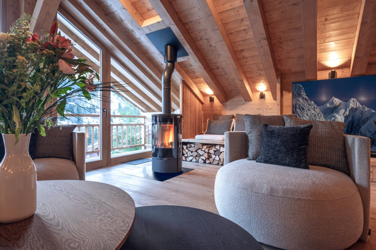 A cozy modern chalet living room with a central black wood-burning stove, round beige poufs, a round wooden coffee table with a white vase of flowers, and a large mountain landscape print on the wall.