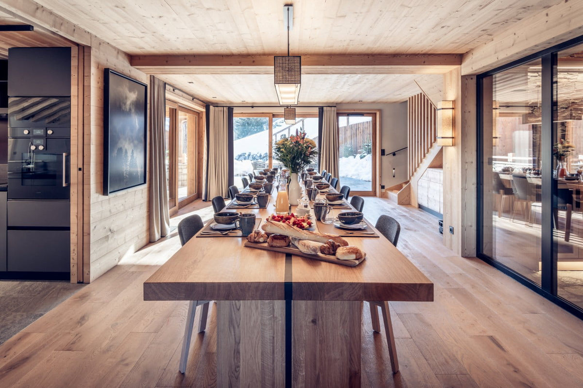 A long wooden dining table set with plates and glassware in a modern chalet-style dining room with wood walls, large windows, and a staircase in the background.