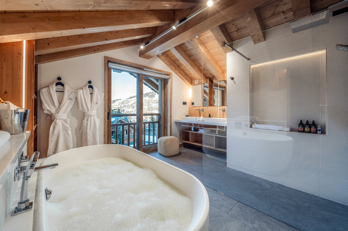 Luxury spa bathroom featuring a large white freestanding bathtub filled with bubbly water, rustic wooden ceiling beams, two white robes hanging on the wall, a modern vanity, and a large window overlooking a snowy mountain landscape.