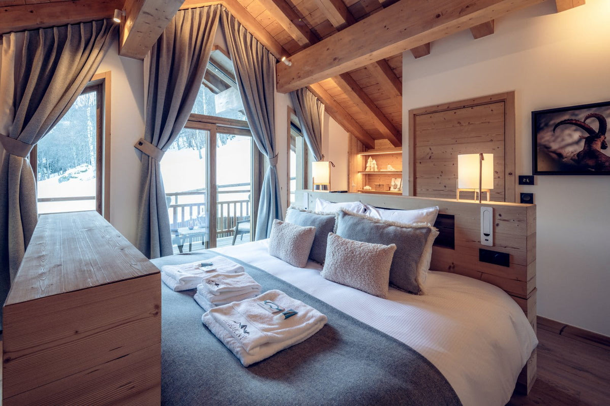 A cozy mountain-style bedroom with a neatly made bed, white linens, gray pillows, wooden ceiling beams, large windows with gray curtains, and a wooden chest in the foreground.