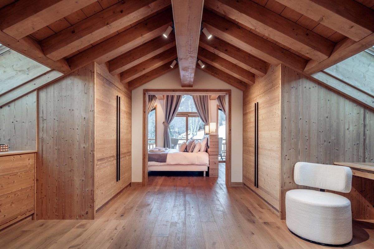 A modern rustic bedroom interior with light wood paneled walls and ceiling, exposed wooden beams, a white upholstered chair in the foreground, and a bed visible through an open doorway in the distance.