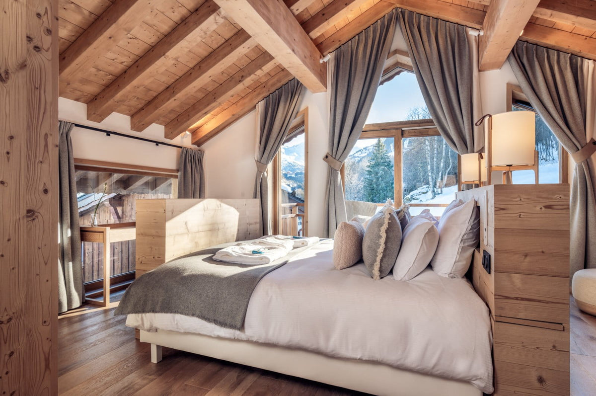 A cozy mountain chalet bedroom featuring a large bed with white linens and gray accents, set under a sloped wooden ceiling with exposed beams. Large windows with gray curtains offer a view of snowy outdoor mountains, and warm bedside lamps sit on wooden nightstands.