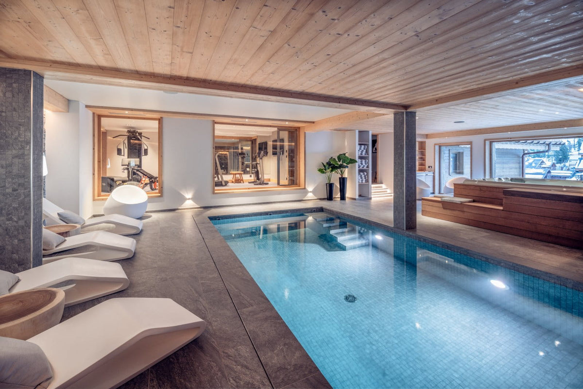 An indoor resort-style swimming pool with blue water, dark stone deck flooring, white lounge chairs along one side, a light wood ceiling, and a modern reception or bar area in the background.