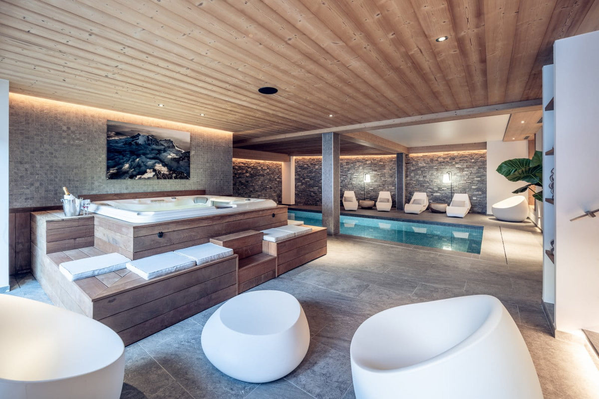 Luxury indoor resort spa and pool area with a wooden hot tub, modern white lounge chairs, a small pool, stone walls, and a wood-paneled ceiling.