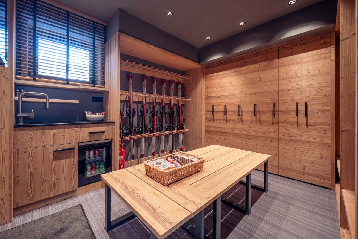 A modern ski lodge or sauna room with light wood cabinetry, a wooden table in the center, a ski rack with skis on the wall, and a small sink area under a window with blinds.