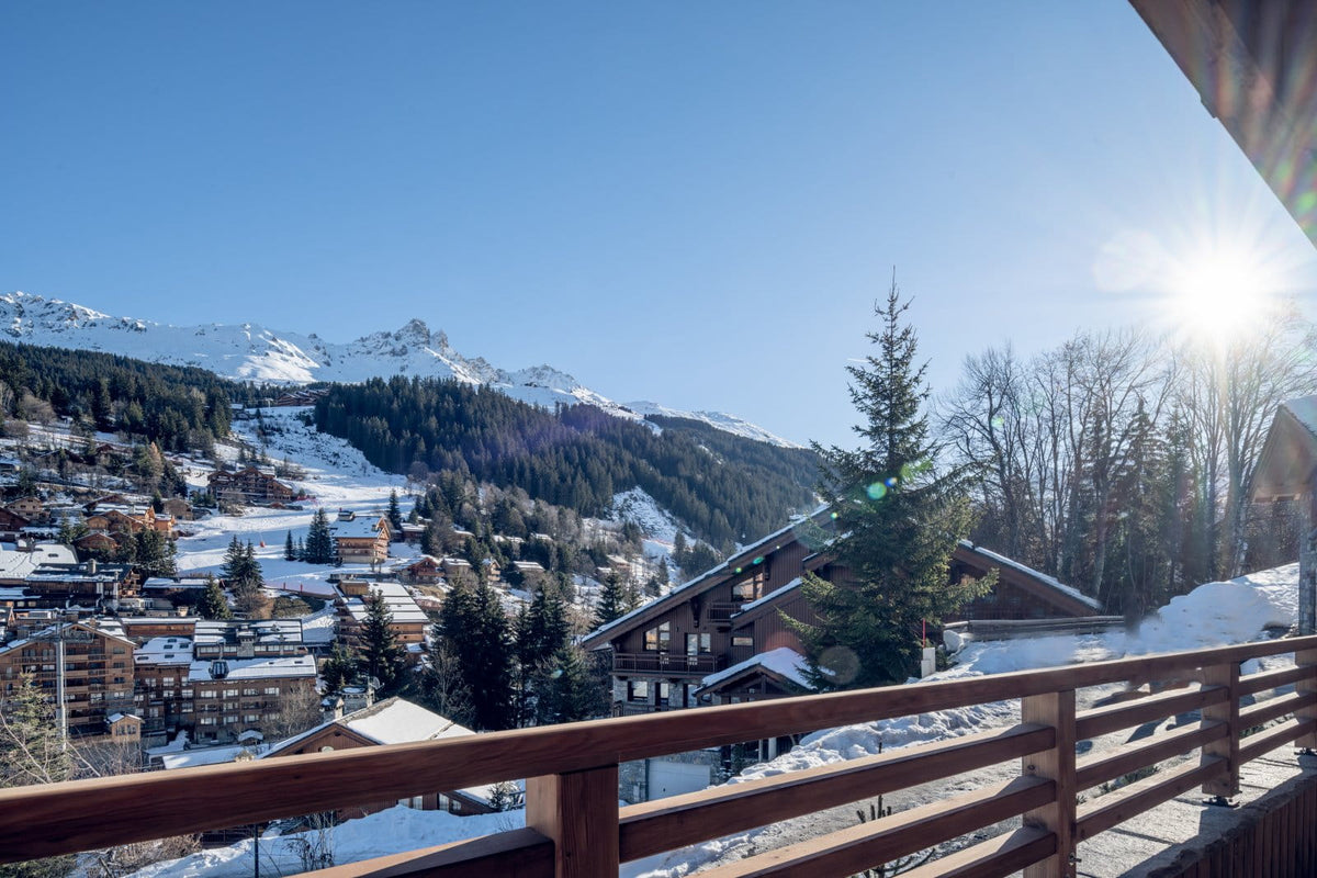 A scenic winter mountain village with snow-covered slopes, alpine chalets, evergreen trees, and a clear blue sky, viewed from a wooden balcony with the sun shining brightly in the upper right.