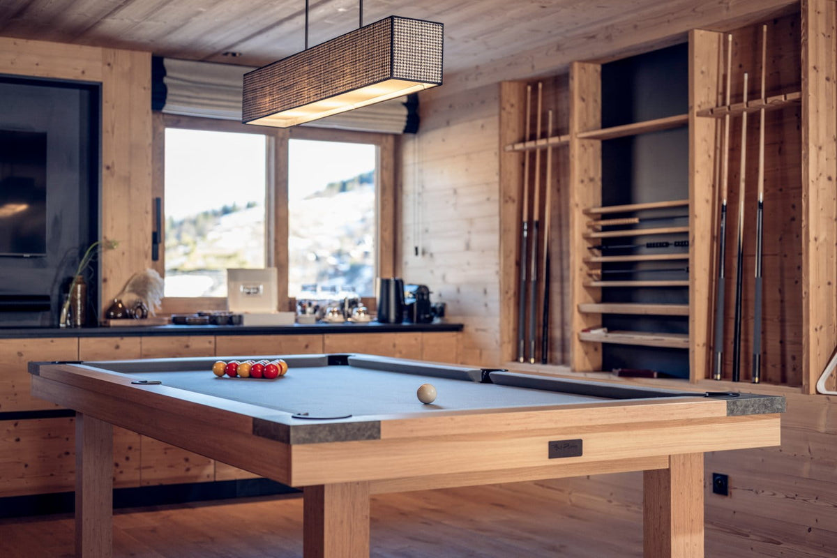 A wooden pool table in a rustic mountain chalet with wood-paneled walls, a hanging light fixture, and cue racks filled with cue sticks.