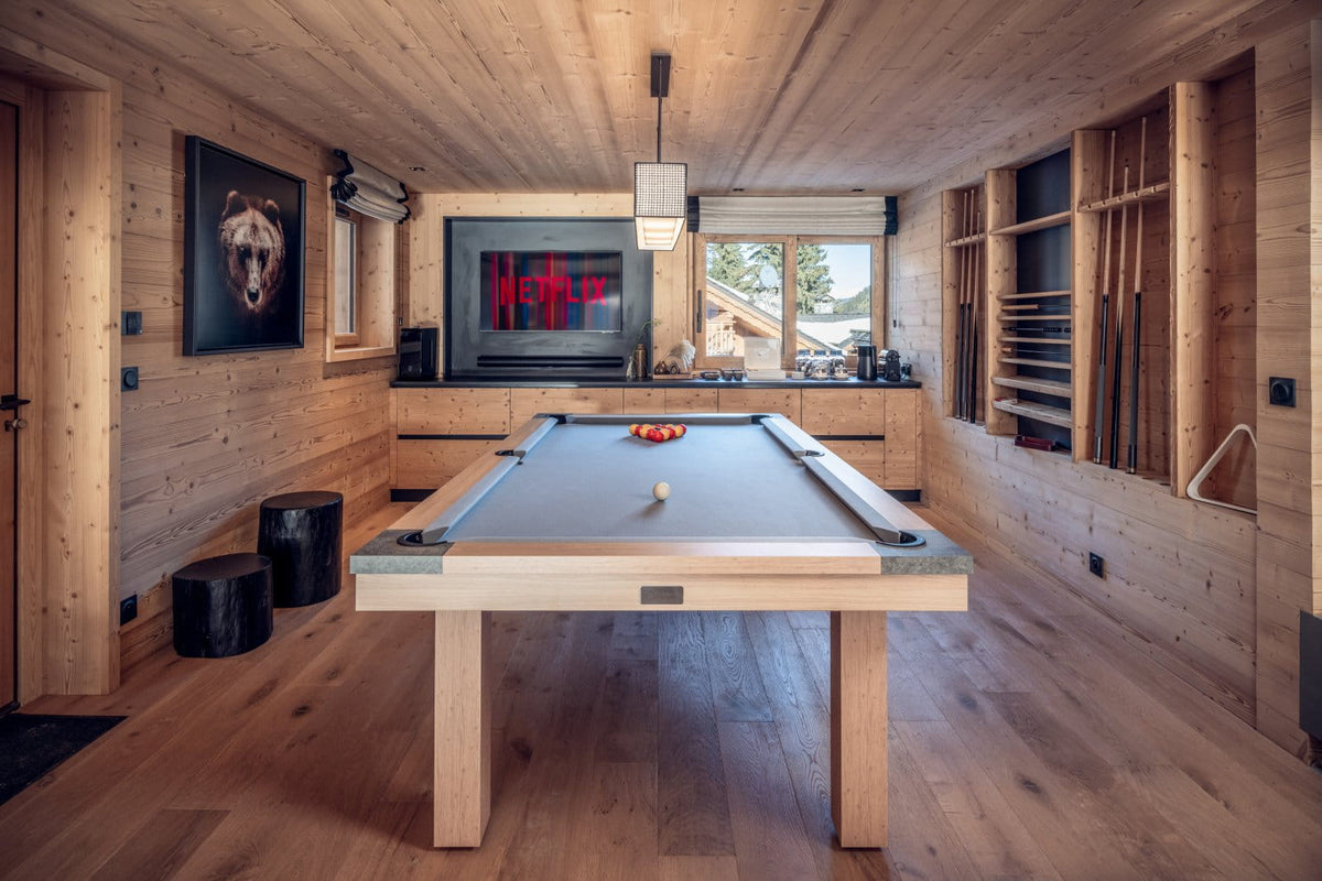 A modern wooden billiards table with a gray felt playing surface is shown in a rustic, wood-paneled alpine-style room with a fireplace and shelves of pool cues.