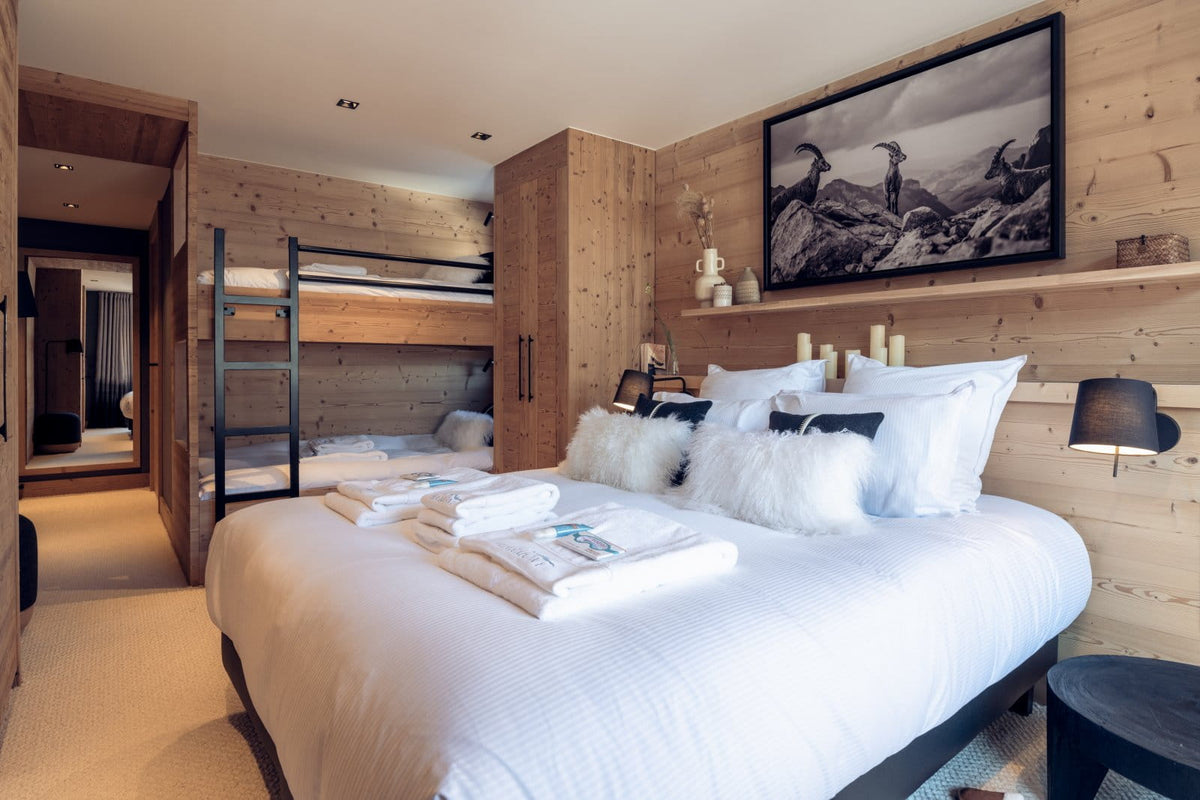A modern mountain-style bedroom with a large bed dressed in white linens and fluffy white pillows, set against a light wood-paneled wall with a black-framed landscape print. A wooden bunk bed and an open doorway to an ensuite bathroom are visible in the background.