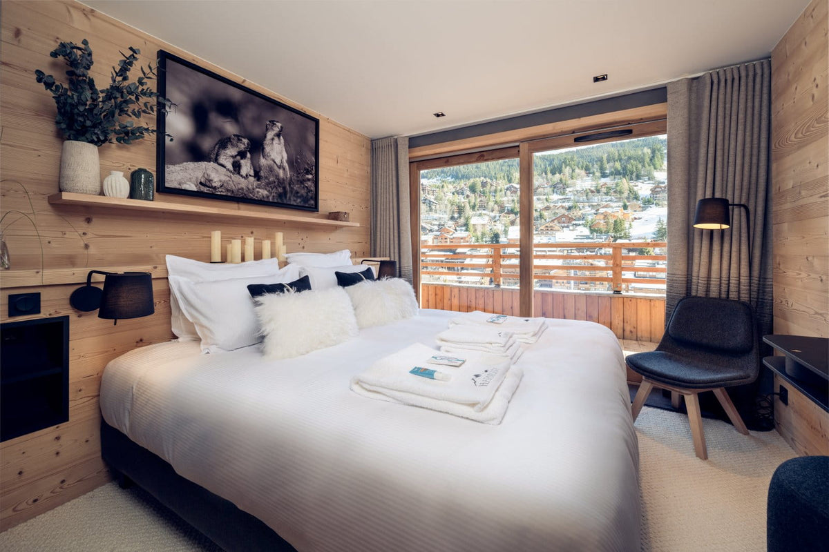 A modern ski chalet bedroom featuring a large bed with white linens, a wooden headboard wall with a framed black-and-white photo, a balcony with a scenic mountain view, and a black accent chair.
