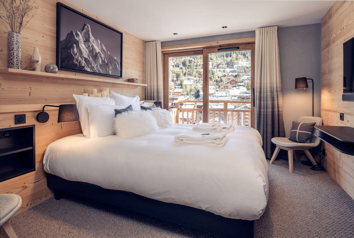 A modern mountain-view bedroom with a large bed dressed in white linens, wood-paneled walls, a framed mountain print, and floor-to-ceiling windows overlooking a ski resort.
