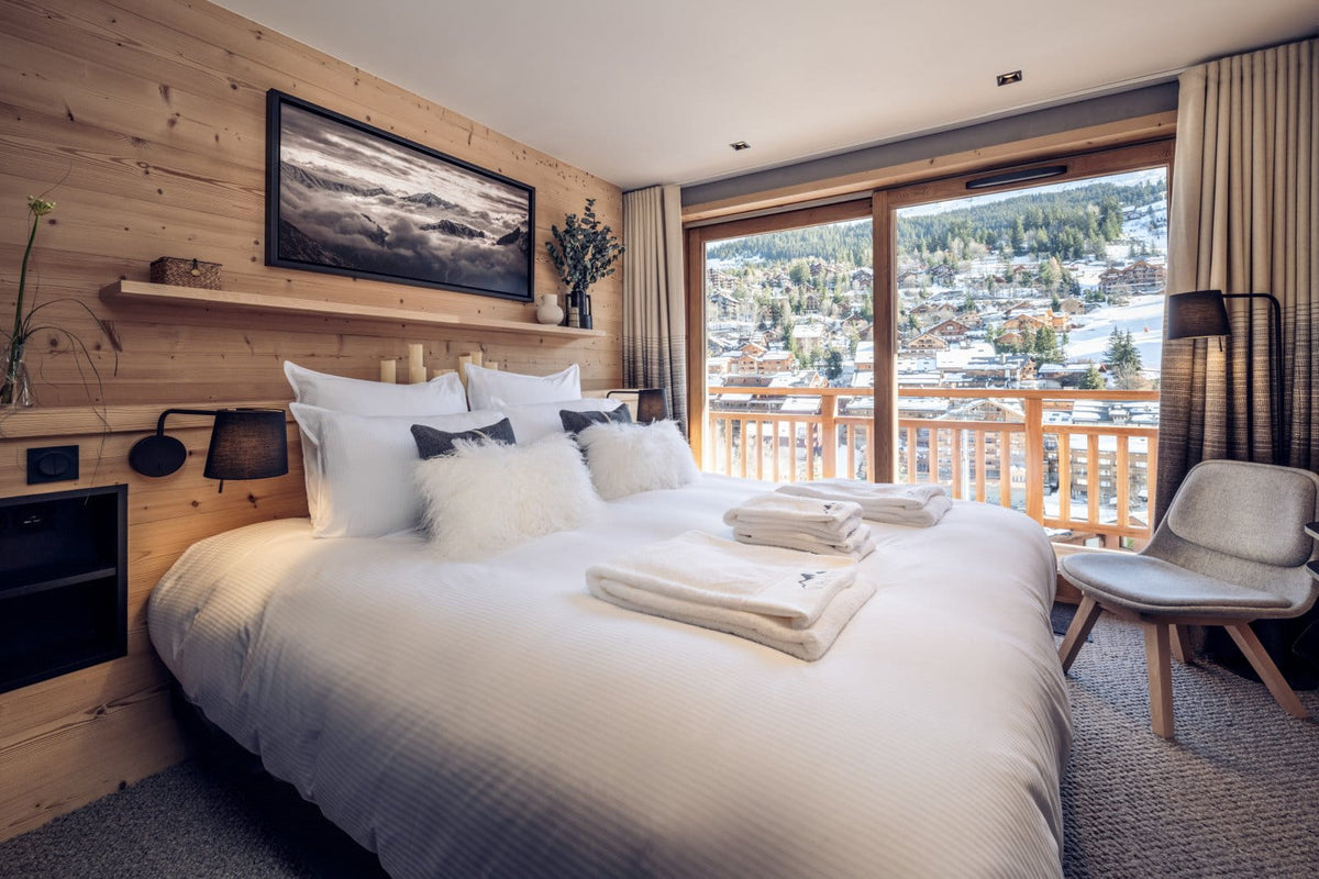 A modern ski chalet bedroom featuring a large bed with white linens, a wooden wall, and a large window overlooking a snowy mountain village.