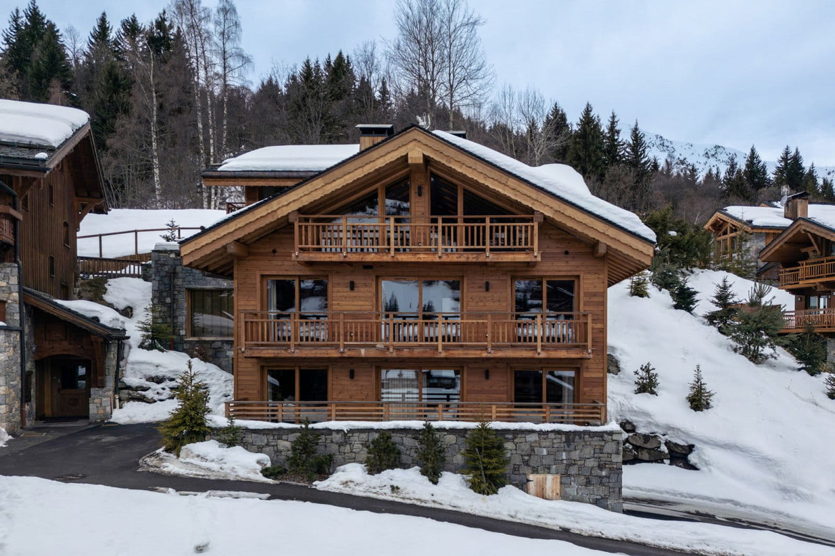 A snow-covered wooden chalet mountain house with large windows and balconies, surrounded by snow and evergreen trees.