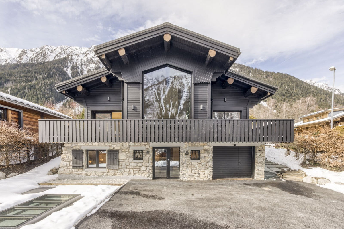 A modern alpine-style chalet with dark gray wood siding, a stone base, large glass windows, a covered balcony, and a driveway leading to a garage, set against snowy mountains and bare winter trees.
