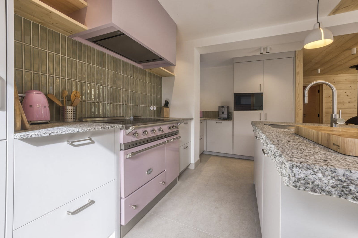 A modern kitchen interior featuring a prominent retro-style pink double-burner gas stove and matching pink upper range hood, set against a green tiled backsplash and white cabinetry.