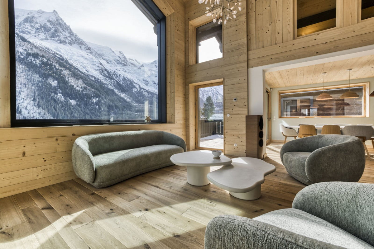 A modern mountain chalet living room with light wood walls and floors, featuring a large window showing a snowy mountain view. The room is furnished with gray upholstered curved sofas, a white sculptural coffee table, and a dining area in the background.