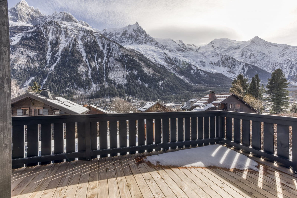 A wooden deck with a dark railing overlooking a snowy mountain village and rugged snow-covered peaks under a bright sky.