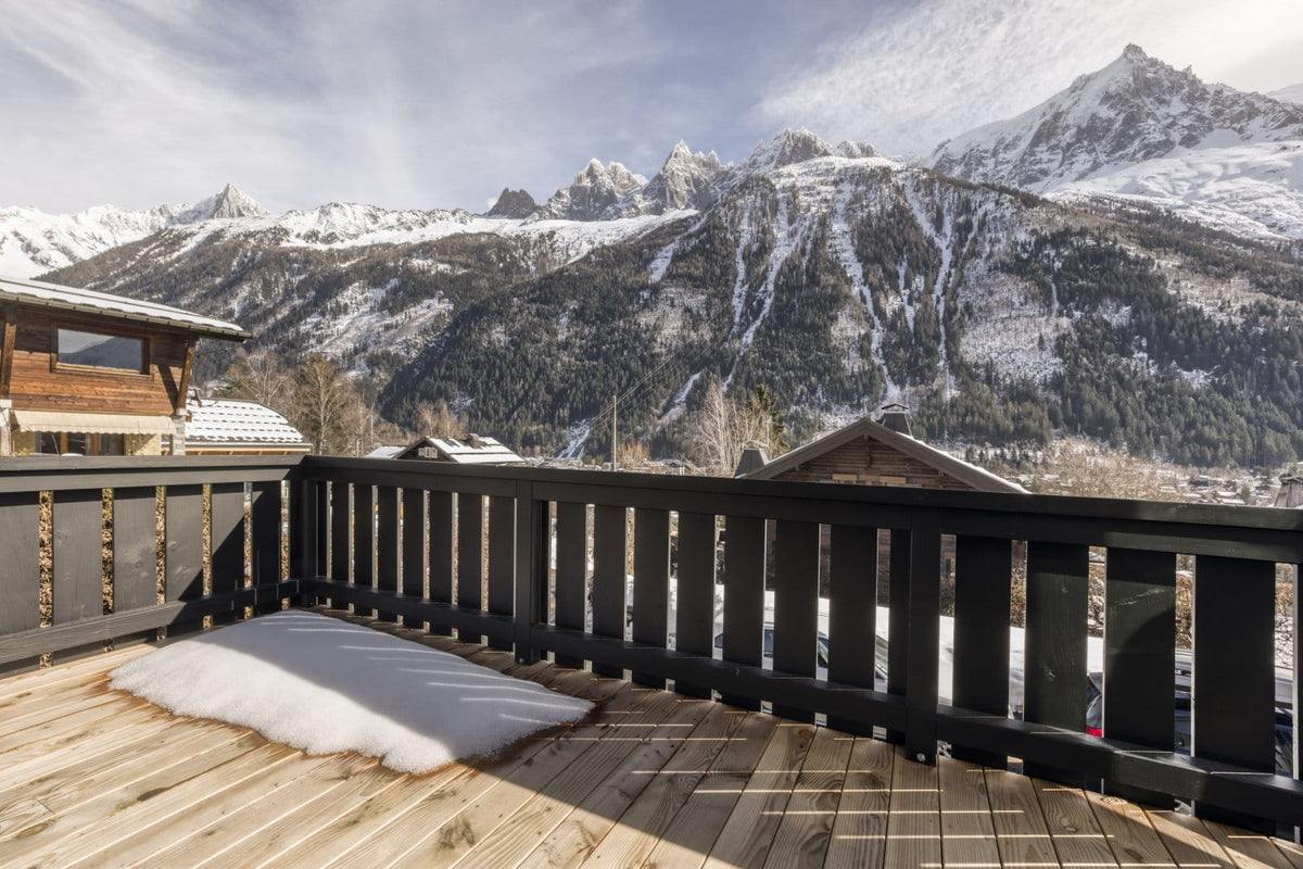 A wooden balcony overlooking a snowy mountain village with snow-covered peaks under a cloudy sky. A white rug sits on the wooden deck, and dark railings frame the foreground.
