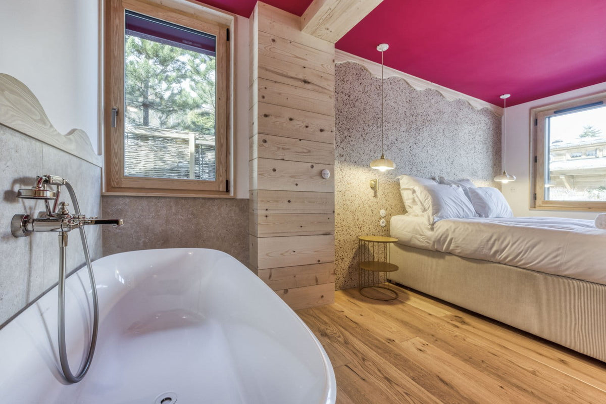 A luxury bedroom and bathroom interior featuring a white freestanding bathtub in the foreground, a bed with white linens, light wood walls and flooring, and a bold magenta-pink ceiling.
