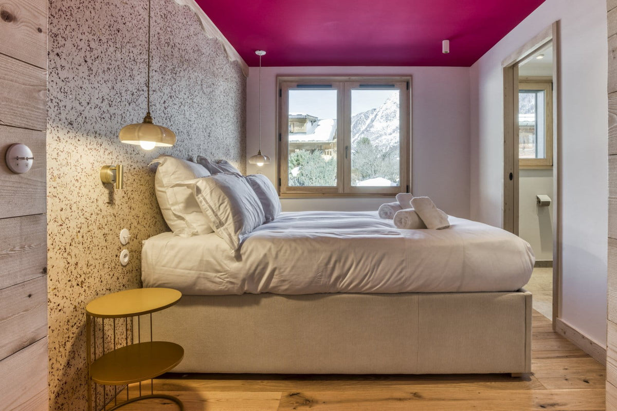 A modern bedroom featuring a low bed with white bedding, a textured speckled wall, light wood paneling, a bright magenta ceiling, and a window showing a snowy mountain view.