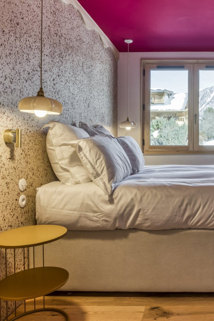 A modern bedroom featuring a low bed with white bedding, a textured speckled wall, light wood paneling, a bright magenta ceiling, and a window showing a snowy mountain view.
