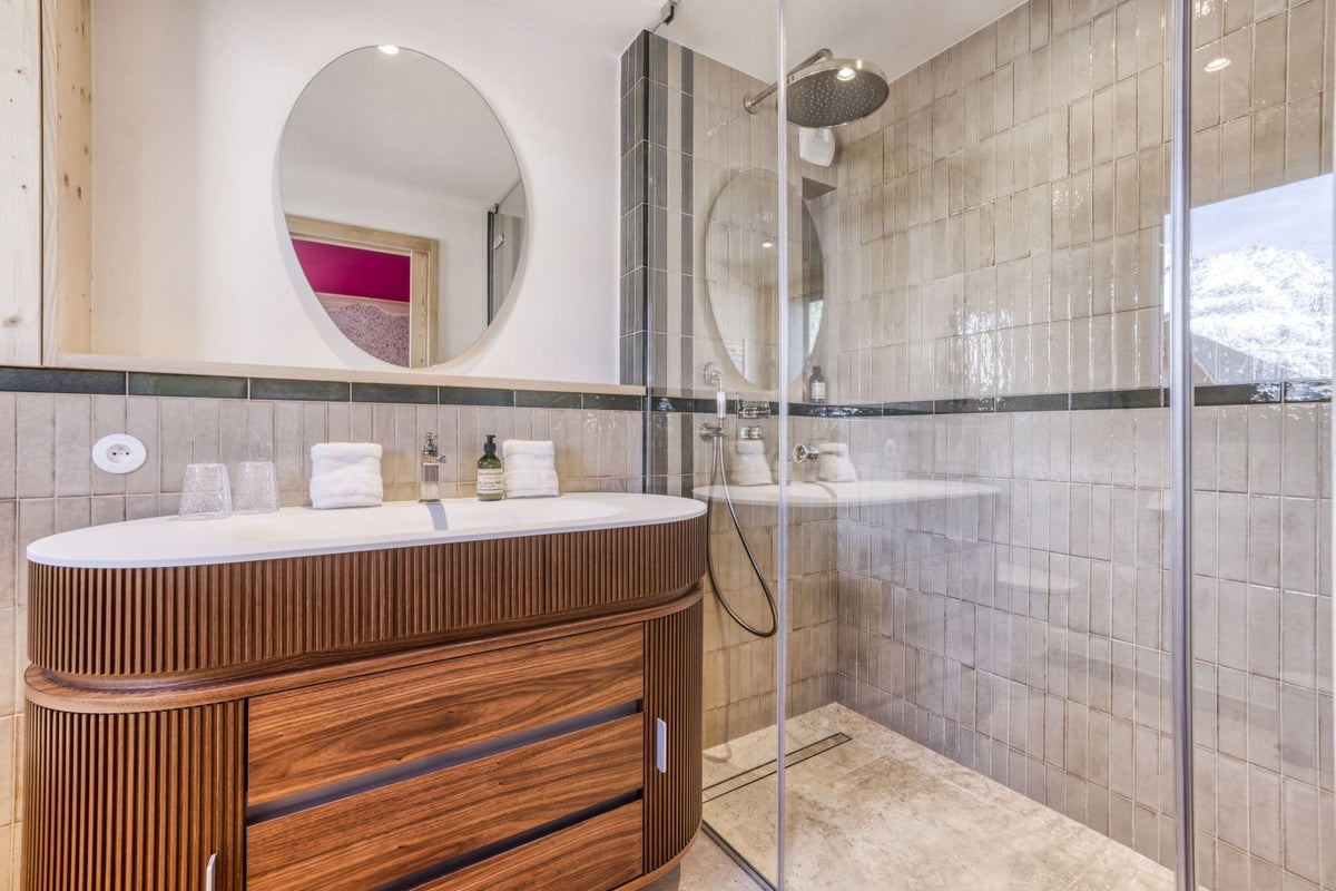 Modern bathroom interior featuring a curved wooden vanity with a white countertop, a round wall mirror, and a glass-enclosed shower area with beige tiled walls.