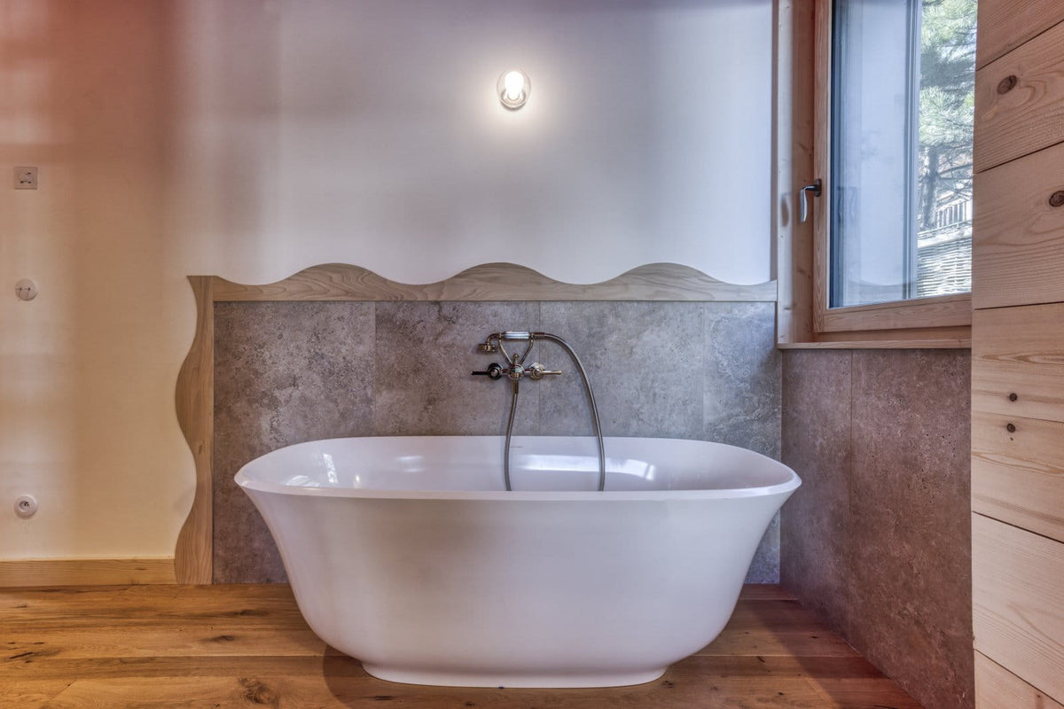 A freestanding oval white bathtub with a chrome faucet and handheld shower attachment, set in a rustic bathroom with wood flooring, a stone backsplash, and a window.