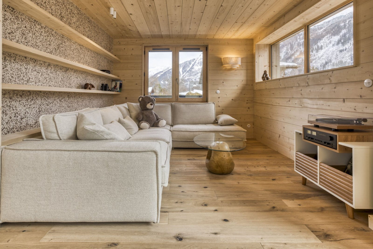 A modern minimalist living room with light wood paneled walls and ceiling, featuring a cream-colored sectional sofa, a small round glass coffee table with a gold base, and a wooden media console with audio equipment. A teddy bear sits on the sofa, and windows in the background show a snowy mountain view.