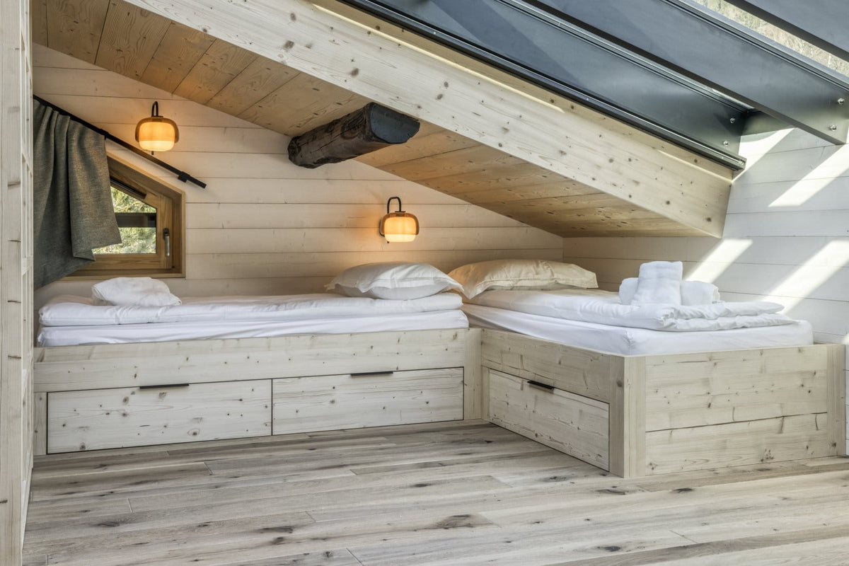 A cozy attic bedroom with two light wood platform beds with white bedding, built into a slanted wooden ceiling with a log accent and a small window.