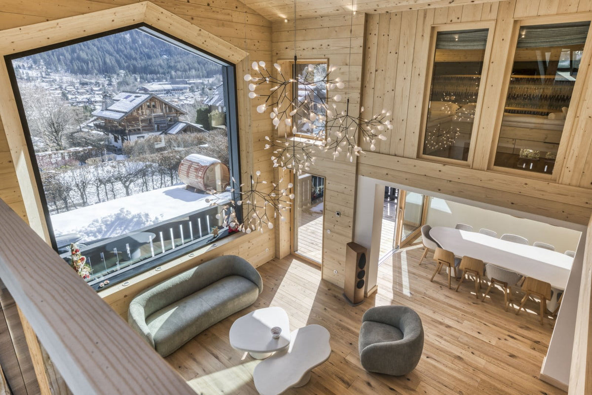 A high-ceilinged wooden chalet interior with a large floor-to-ceiling window overlooking a snowy mountain village. The space features light wood walls and floors, a modern chandelier, a curved gray sofa, a gray armchair, white sculptural side tables, and a dining area in the background.