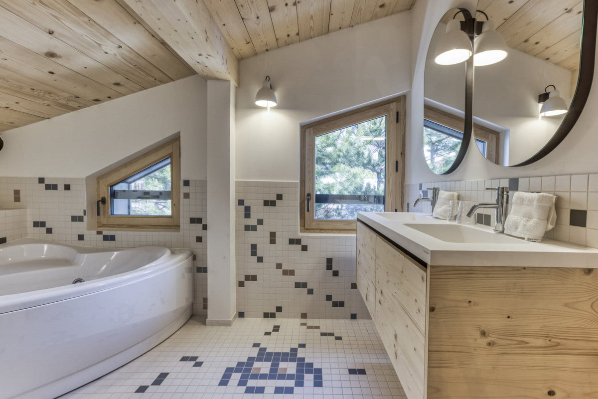 A rustic mountain-style bathroom featuring a white freestanding tub, a light wood vanity with a white countertop, a large round mirror, and blue-and-white mosaic tile accents on the walls and floor.