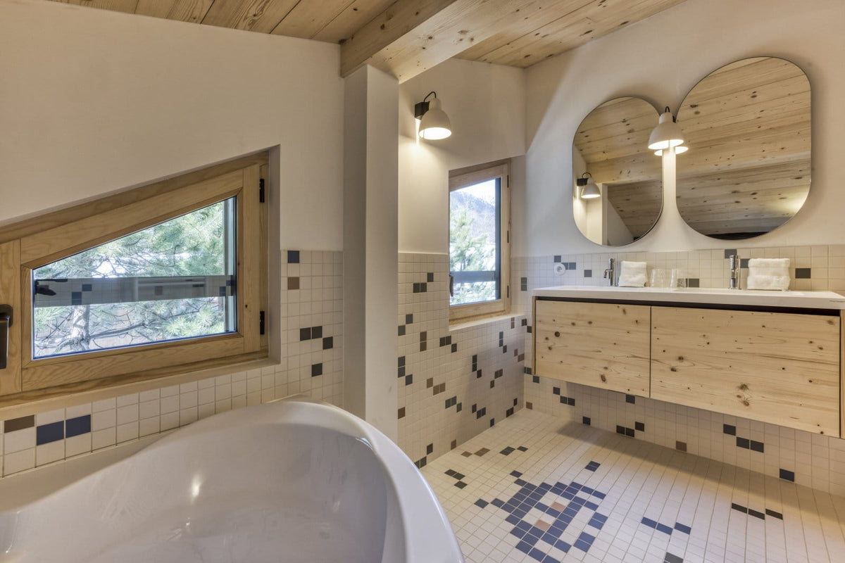 A modern bathroom interior with a freestanding white bathtub in the foreground, light wood ceiling beams, a floating double-sink vanity with two large oval mirrors, and tiled walls and floor with black-and-white mosaic accents.