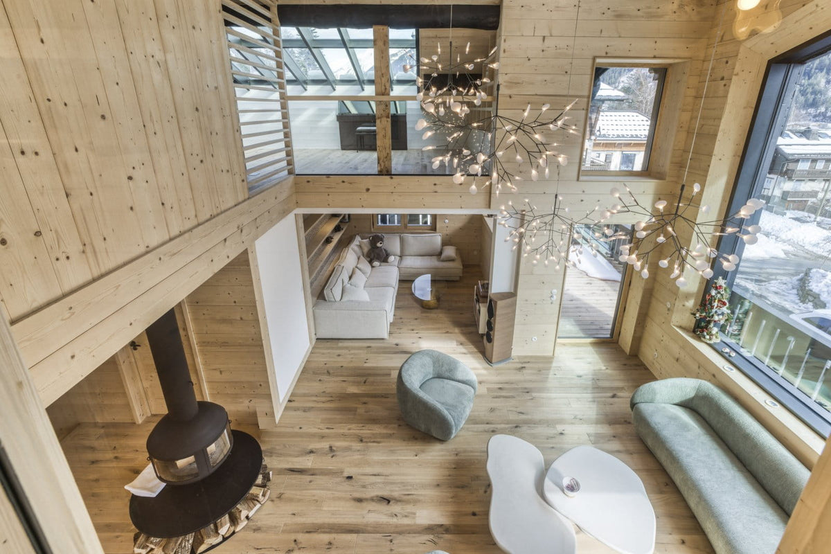 A high-angle view of a modern, rustic chalet living room with light wood walls and floors, featuring a black wood-burning stove, white sofas, a gray armchair, and large windows looking out onto a snowy outdoor scene.