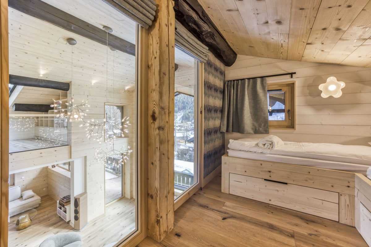 A rustic mountain chalet bedroom featuring light natural wood paneling on the walls and ceiling, a built-in wooden storage bed with white bedding, a flower-shaped wall light, and large glass doors looking out onto a snowy outdoor landscape.