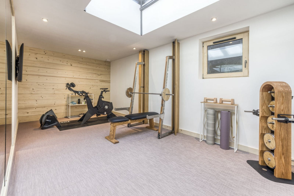 A home gym interior with multiple fitness pieces of equipment, including a weight bench, barbell rack, wooden exercise wall, and a wooden weight stack, set against a light wood paneled wall and white walls with a skylight.