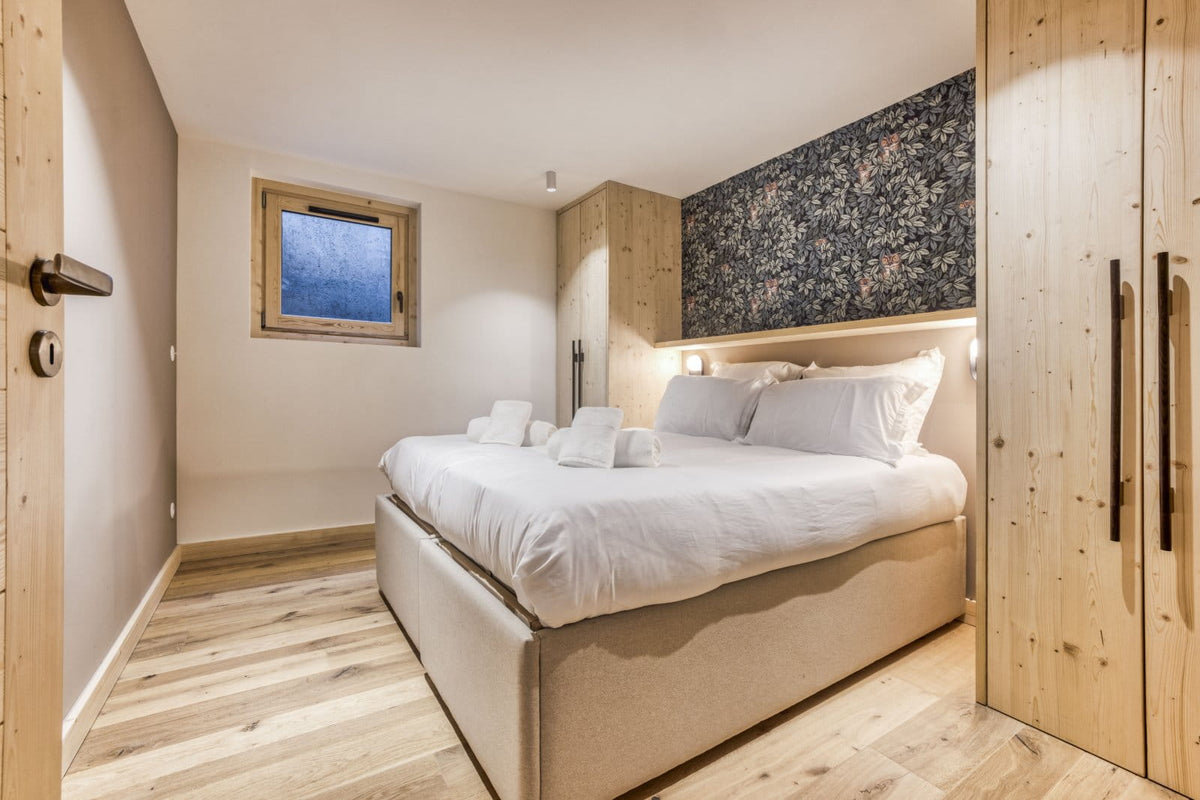 A modern bedroom featuring a low-profile bed with white bedding, light wood flooring, a floral-patterned accent wall, and light wood cabinetry.