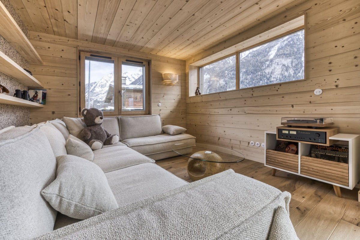 A cozy modern chalet living room with light beige sectional sofas, a glass coffee table, and a wooden media cabinet, set against wood-paneled walls with views of snowy mountains through large windows.