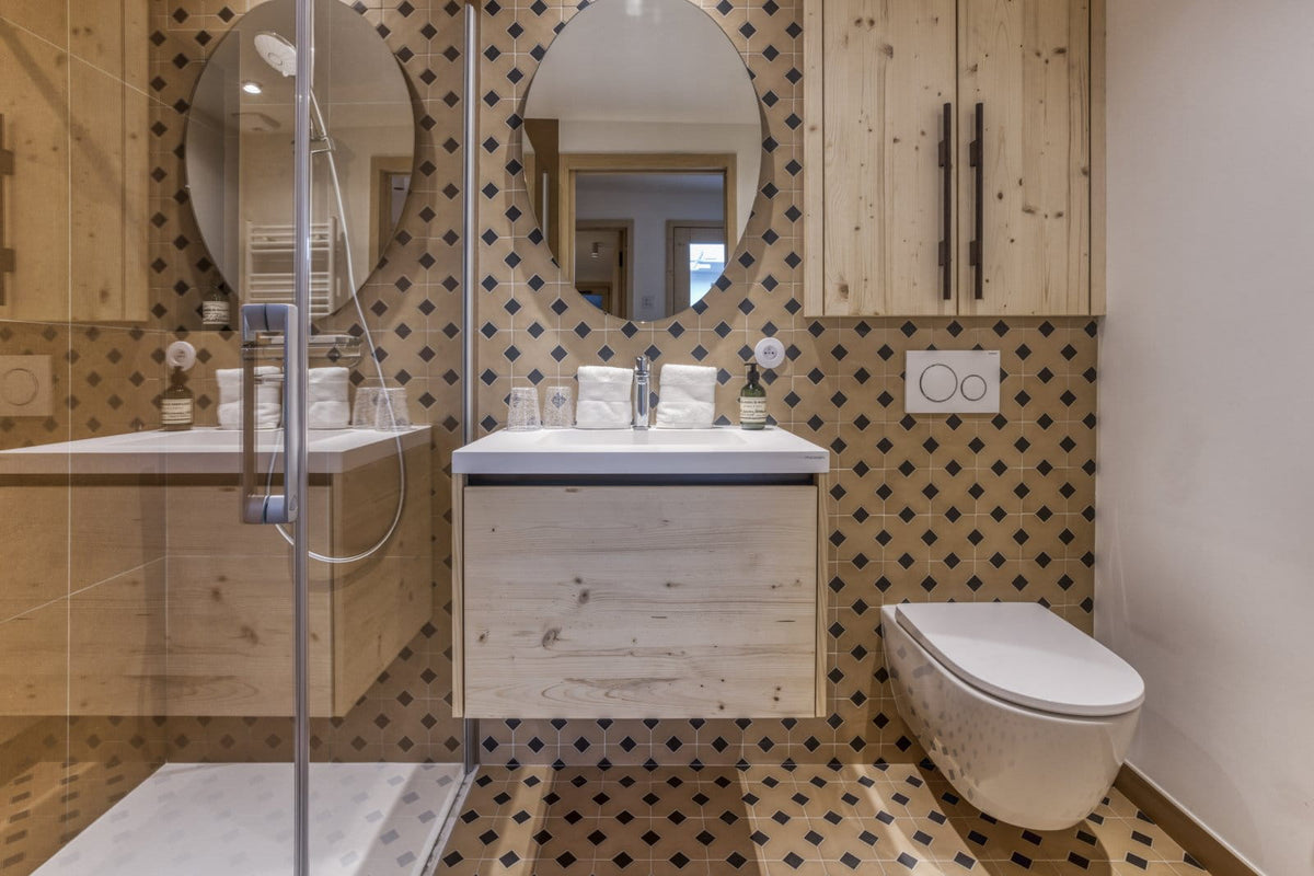 A modern bathroom interior with a floating light wood vanity, white sink, round mirrors, patterned brown and beige wall and floor tiles, a walk-in shower area with glass, and a white wall-mounted toilet.