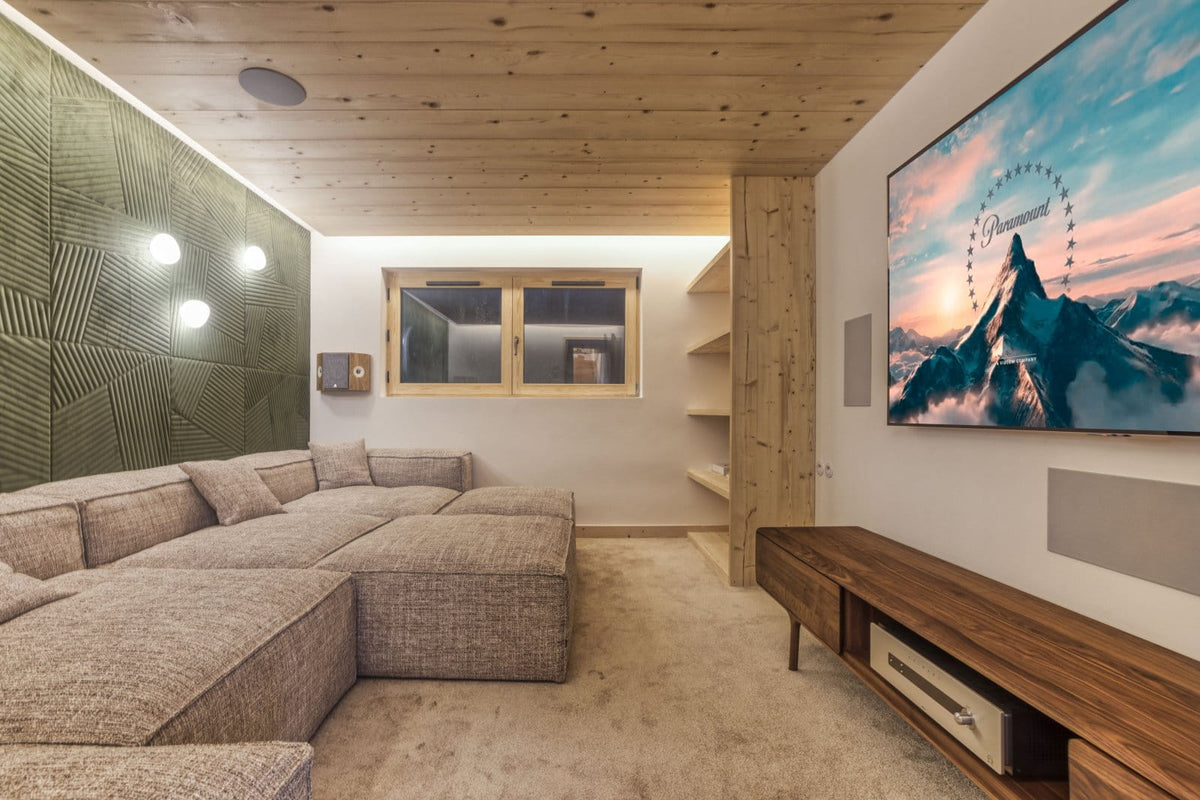 A modern mountain-themed home theater or living room featuring a large sectional sofa, a wooden media console, a wall-mounted TV displaying a mountain landscape, and a textured green accent wall.