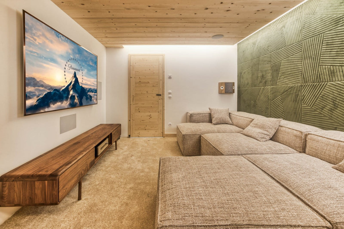 A modern home theater or lounge room featuring a large wall-mounted TV displaying a mountain sunset, a low wooden media console, a beige sectional sofa, a textured green accent wall, and a light wood ceiling.