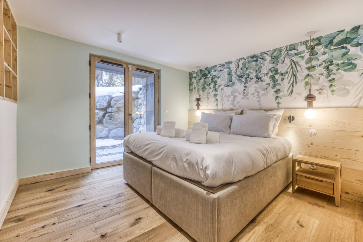A modern bedroom featuring a large bed with white linens and gray pillows, set against a light wood headboard and a green botanical wall mural. The room has light wood flooring, a wooden nightstand, and a glass door leading to an outdoor stone patio.