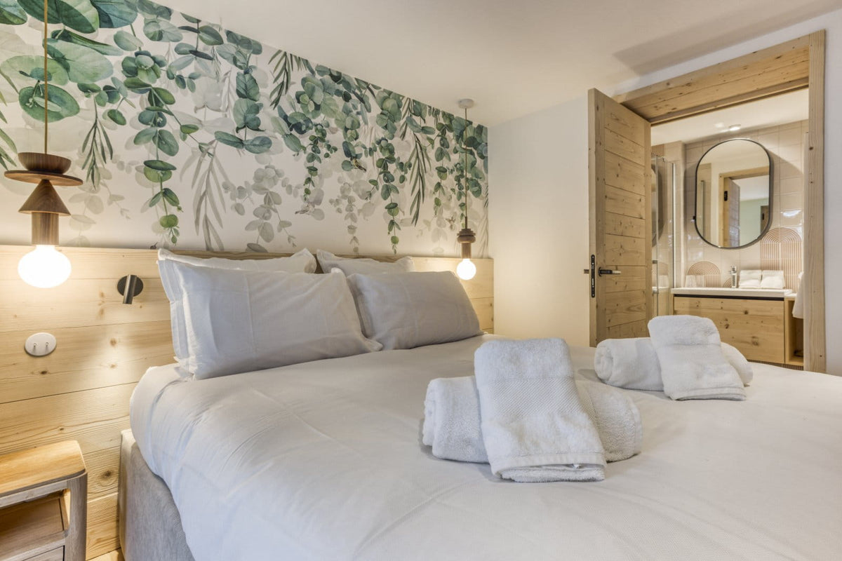 A modern bedroom featuring a large bed with white linens, folded towels, a light wood headboard and nightstand, and a decorative green botanical wall mural behind the bed. An open wooden door reveals a bathroom in the background.