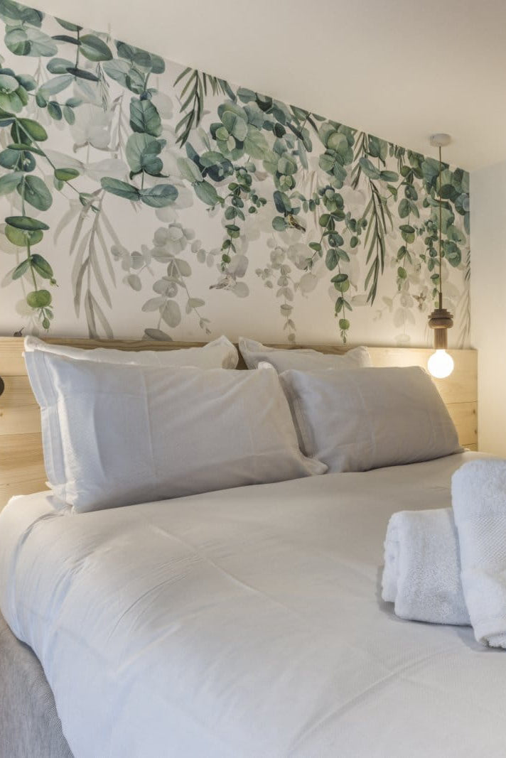 A modern bedroom featuring a large bed with white linens, folded towels, a light wood headboard and nightstand, and a decorative green botanical wall mural behind the bed. An open wooden door reveals a bathroom in the background.