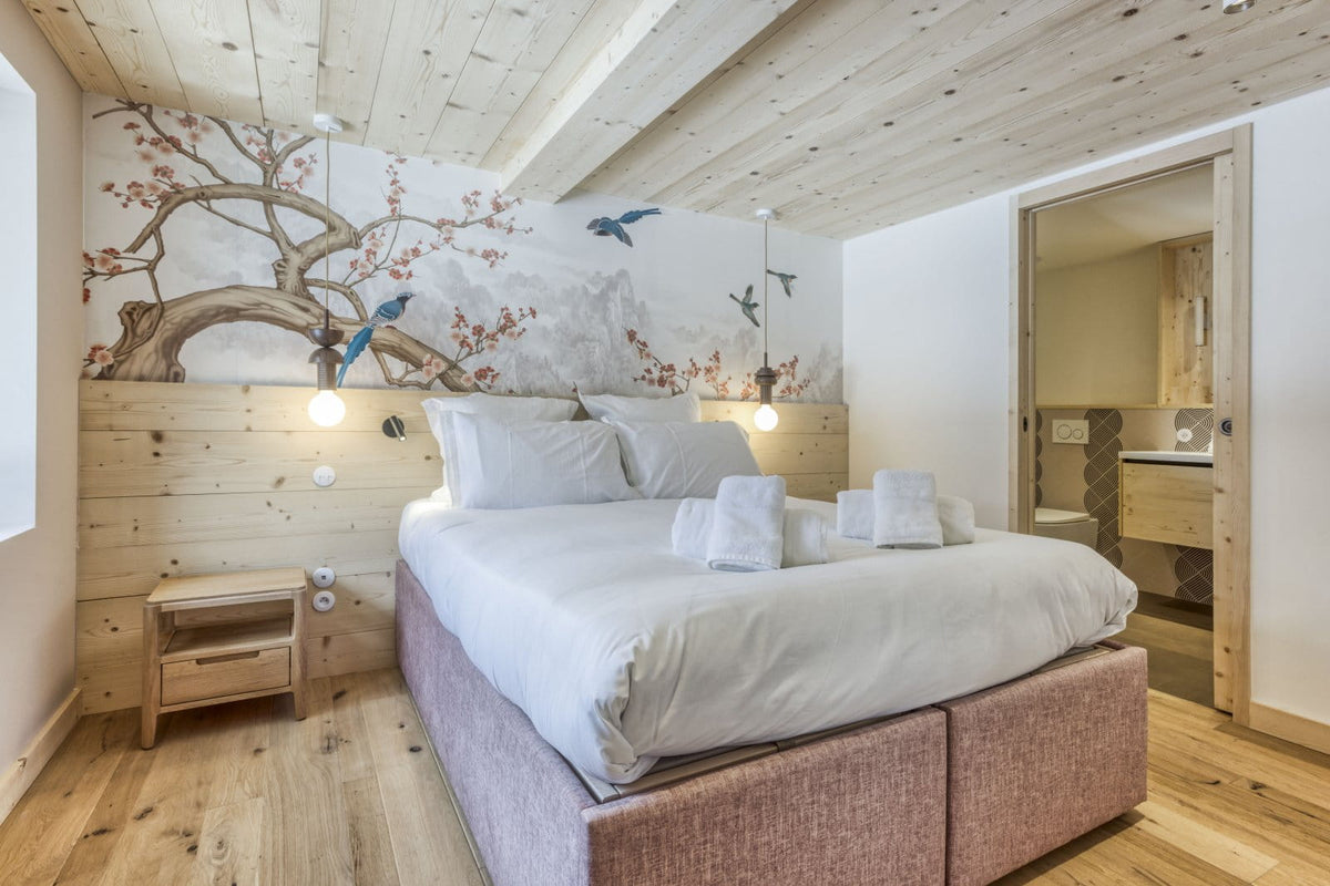 A modern bedroom featuring a large bed with white linens and a textured mauve-pink upholstered base, set against a light wood-paneled wall with a large tree mural and birds. The room has light wood floors and ceiling, a small wooden nightstand, and an open doorway revealing a bathroom.