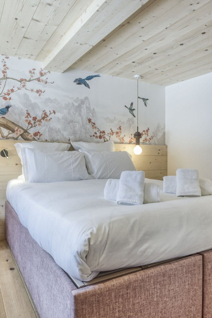 A modern bedroom featuring a large bed with white linens and a textured mauve-pink upholstered base, set against a light wood-paneled wall with a large tree mural and birds. The room has light wood floors and ceiling, a small wooden nightstand, and an open doorway revealing a bathroom.