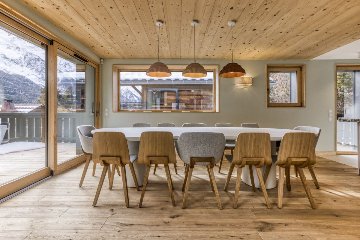 A modern dining room with a long oval white table surrounded by light wood and gray upholstered chairs, set in a rustic mountain chalet with wood-paneled ceilings and large windows overlooking snowy mountains.
