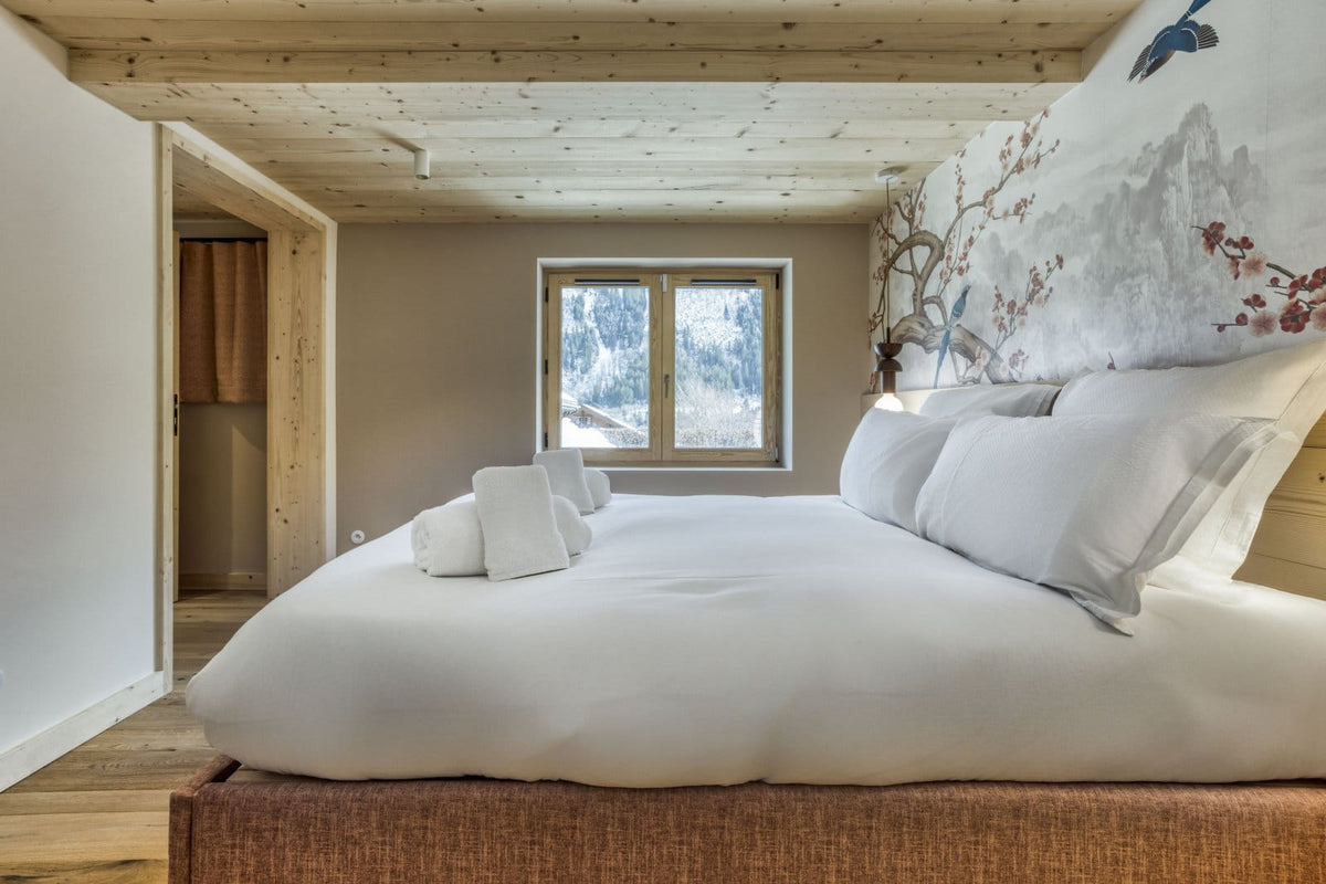 A cozy bedroom interior featuring a neatly made bed with white bedding, a large decorative wall mural with birds and mountains, a window looking out onto a snowy landscape, and light wood ceiling and floor.