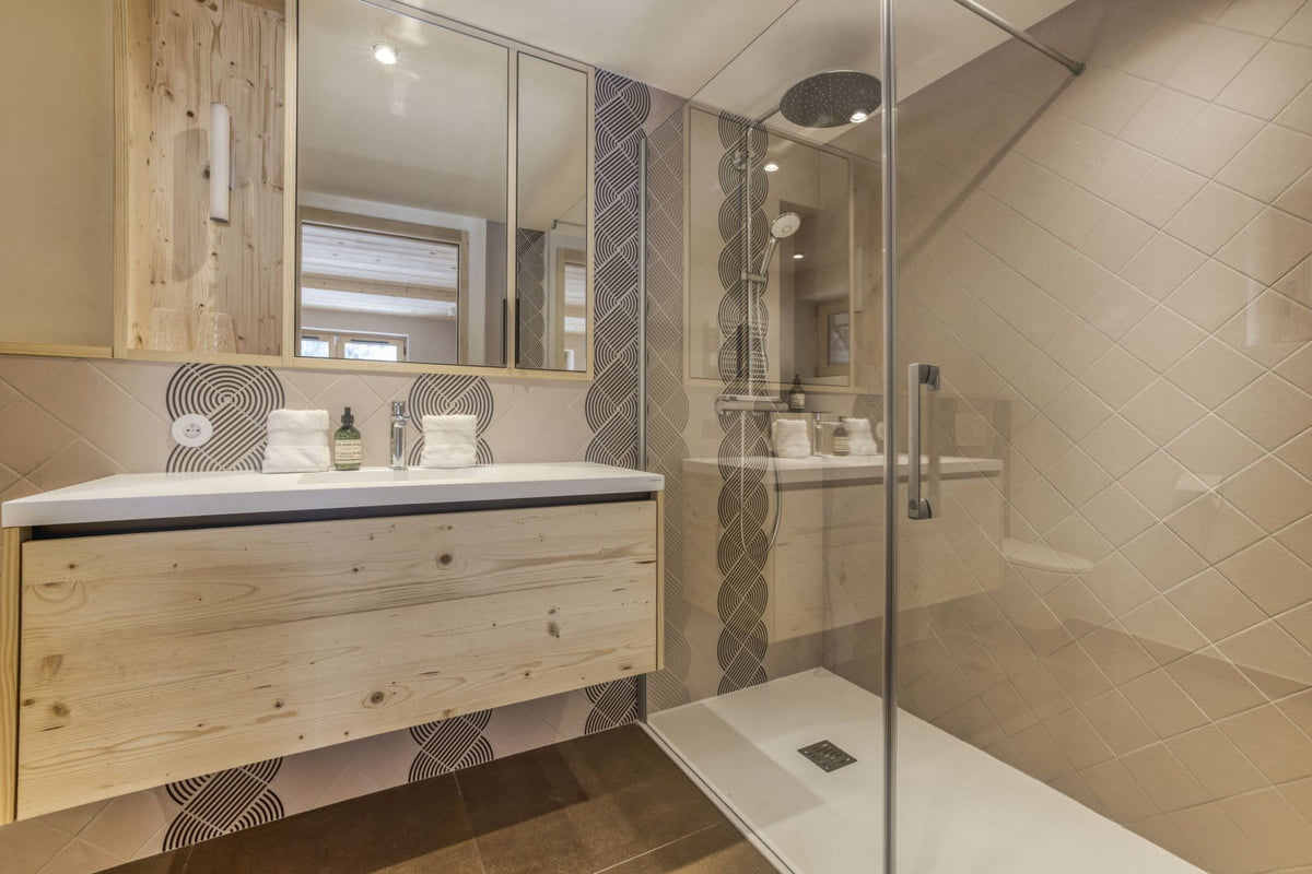 Modern bathroom interior featuring a floating light wood vanity with a white sink countertop, large mirrors, a glass shower enclosure, and beige tiled walls and floor.