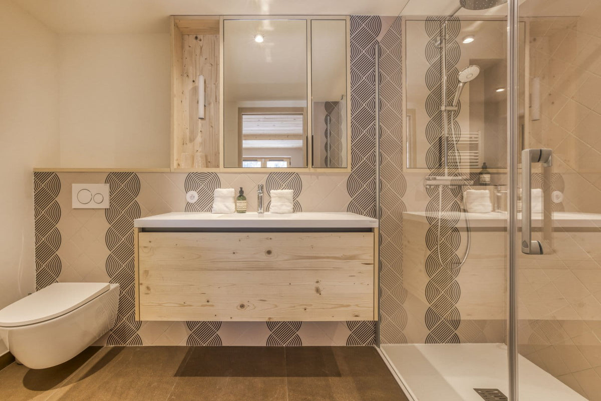 A modern bathroom interior featuring a light wood floating vanity with a white sink, a wall-mounted toilet, a glass shower enclosure, and patterned gray and beige tile accents.