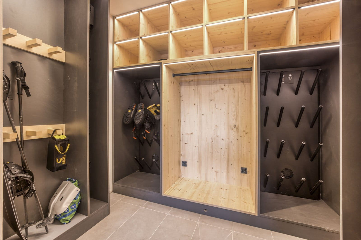 A modern ski and snowboard storage closet with light wood cubby shelves, a central hanging frame, black peg walls for skis and snowboards, and ski equipment stored on the left against a dark gray wall.